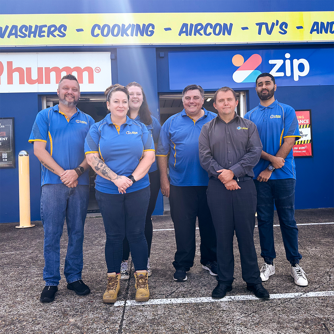 Staff in blue uniforms in front of Bi-Rite Home & Save On Appliances storefront displaying various appliance ads and open 7 days sign.